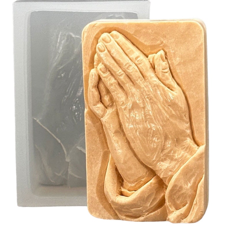 May include: A silicone mold for making soap or other crafts. The mold is shaped like two hands clasped in prayer. The mold is light brown in color and has a smooth, detailed surface.