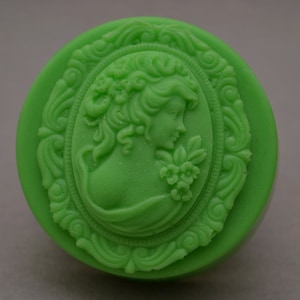 Pode incluir: Um sabonete verde redondo com um design em relevo de um rosto de mulher em uma moldura oval. A moldura é decorada com detalhes intrincados de rolagem.