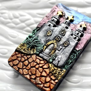 Könnte beinhalten: Eine silber- und goldfarbene Keramikfliese mit einem Reliefbild einer Kirche mit einem Kreuz auf der Spitze. Die Kirche ist von einer grün- und goldfarbenen Landschaft umgeben. Die Fliese befindet sich auf einem weißen Hintergrund.