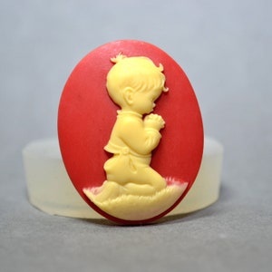 May include: A red oval cameo with a raised image of a child kneeling in prayer. The child is wearing a white shirt and pants and has blonde hair.