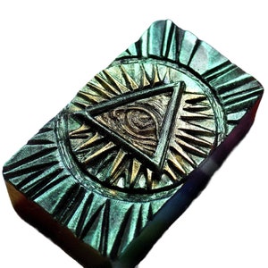 Può includere: Una saponetta rettangolare verde e oro con un design di occhio onniveggente. L'occhio è all'interno di un triangolo, che è all'interno di un cerchio con un motivo a stella.