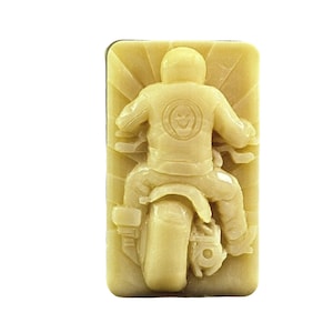 May include: A beige soap bar featuring a raised design of a person riding a motorcycle. The motorcycle has a large rear tyre and the person is wearing a jacket with a skull design on the back.