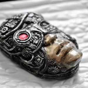 Puede incluir: Una escultura de rostro de robot de plata y oro con un ojo rojo. La escultura está detallada con engranajes y patrones intrincados.