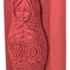May include: A red, rectangular stamp featuring a detailed carving of a Matryoshka doll. The doll's face is visible, and the design includes floral patterns and a headscarf. The stamp is a solid block with a textured surface.