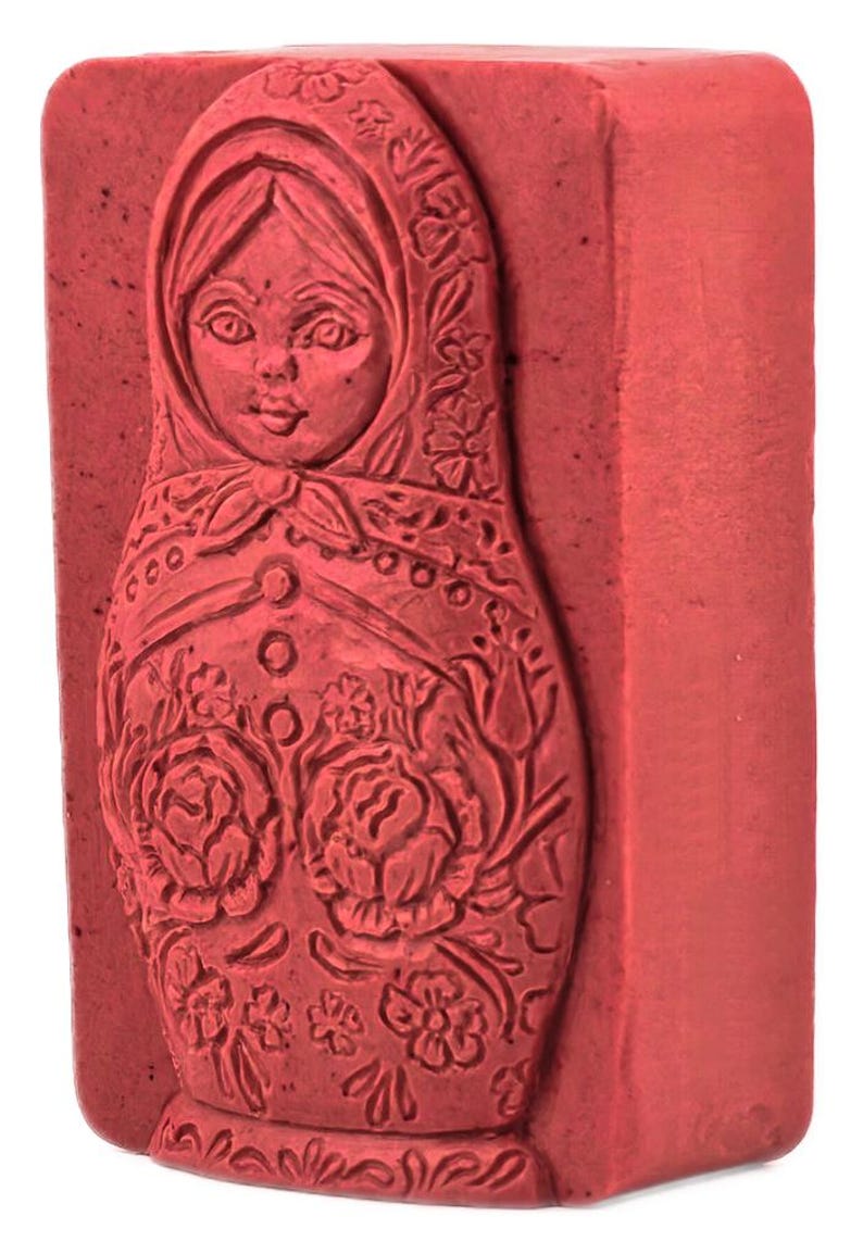 May include: A red, rectangular stamp featuring a detailed carving of a Matryoshka doll. The doll's face is visible, and the design includes floral patterns and a headscarf. The stamp is a solid block with a textured surface.
