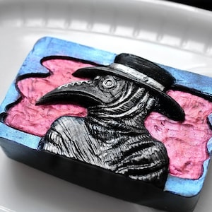 May include: A black and silver plague doctor soap bar with a blue and pink background. The soap features a detailed carving of a plague doctor wearing a long coat and a beak mask.
