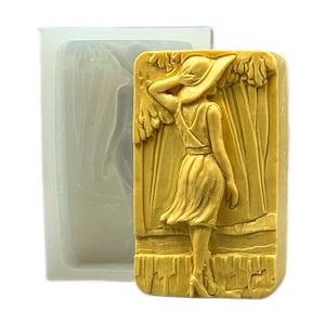 Könnte beinhalten: Ein rechteckiger, hellgelber Seifenblock mit einem detaillierten Relief einer Frau in Kleid und Hut, die neben einem stilisierten Baum steht. Eine transparente, rechteckige Silikonform befindet sich neben der Seife. Die Seife ist ein dekorativer Artikel.