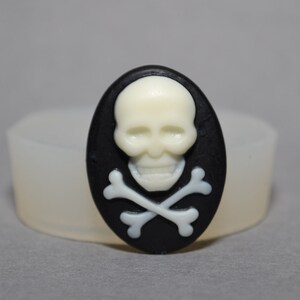 May include: A black oval cameo with a white skull and crossbones design. The skull has a detailed face with eye sockets, a nose, and a mouth.