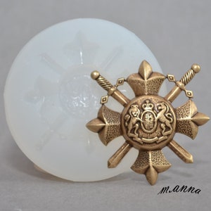 May include: A silicone mold for making a decorative pendant featuring a shield with two crossed swords. The shield has a crown and two lions on it. The mold is white and the pendant is gold.