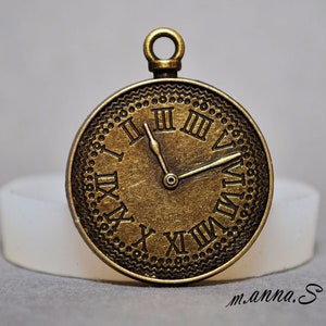 Può includere: Un ciondolo color bronzo a forma di quadrante di orologio con numeri romani e un paio di lancette. Il quadrante dell'orologio ha un bordo decorativo di piccoli punti e una linea ondulata.