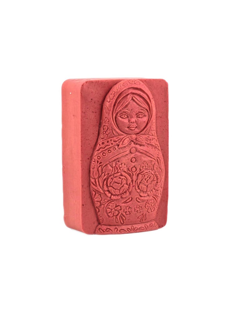 May include: A rectangular, reddish-pink soap bar featuring a detailed relief of a Matryoshka doll. The doll's design includes a headscarf, floral patterns, and a symmetrical arrangement of buttons. The soap is a solid color with a smooth texture.
