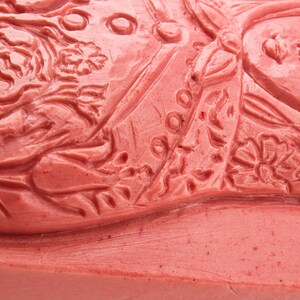 May include: Close-up of a red rubber stamp with a detailed relief design. The stamp features floral and human figure motifs, with intricate patterns and textures. The design is carved into the red rubber surface.