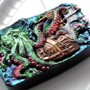 Op de afbeelding: Een donkerblauw, groen en rood harsbeeld van een octopus die een schip aanvalt. Het beeld heeft een metallic, iriserende afwerking.