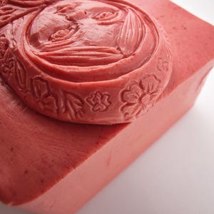 May include: A rectangular bar of pink soap with a carved face design. The face is surrounded by floral and leaf patterns. The soap has a smooth surface and a slightly angled edge, with a light background.