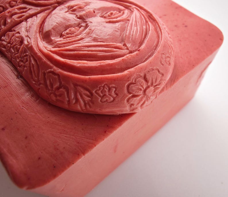May include: A rectangular bar of pink soap with a carved face design. The face is surrounded by floral and leaf patterns. The soap has a smooth surface and a slightly angled edge, with a light background.