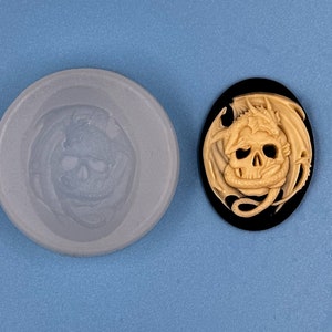 May include: A beige resin cameo featuring a detailed sculpture of a dragon wrapping around a skull. The cameo is set in a black oval frame.