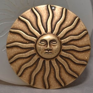 Puede incluir: Un colgante de sol de metal dorado con una cara y rayos. El sol es redondo y tiene una superficie texturizada.