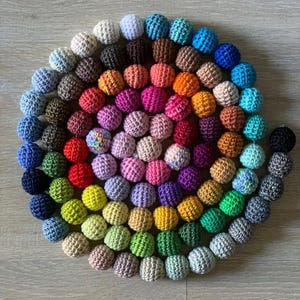 Könnte beinhalten: Eine spiralförmige Anordnung von gehäkelten Kugeln in verschiedenen Farben, darunter Blau, Grün, Gelb, Rosa und Braun. Die Kugeln sind gleichmäßig groß und auf einer hellbraunen Holzoberfläche angeordnet. Die handgefertigten Artikel sind wahrscheinlich für Dekoration oder Basteln gedacht.