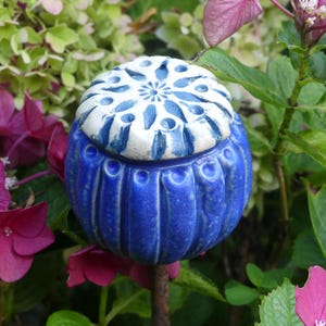 Op de afbeelding: Blauw en wit keramisch tuinornament. Het bolvormige ornament heeft een blauwe basis met verticale groeven en een witte bovenkant met een bloemmotief. Het staat tegen een achtergrond van groen gebladerte en roze bloemen.