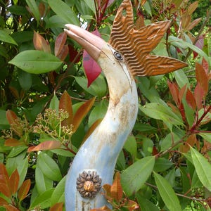 Op de afbeelding: Een keramisch vogelsculptuur met een blauw en wit lichaam, een gouden kroon en een roze snavel. Het beeld staat in een tuin met groene bladeren.