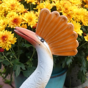 Op de afbeelding: Een wit keramiek tuinbeeld van een kraanvogel met een oranje waaiervormige kam. De kraanvogel staat voor een pot met gele chrysanten.
