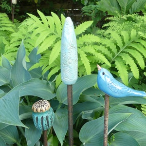 Op de afbeelding: Drie keramische tuinpalen, een in de vorm van een vogel, een in de vorm van een peul en een in de vorm van een hoge, dunne bloem. De vogel is blauw, de peul is groen en bruin, en de bloem is lichtblauw met witte swirls. Alle drie zijn op roestige metalen palen.
