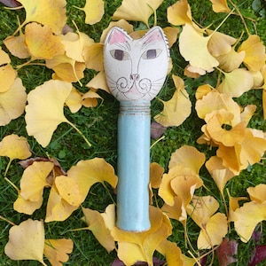 Op de afbeelding: Een wit keramiek kattenhoofd met zwarte ogen en een roze neus, bevestigd aan een lichtblauwe keramische cilinder. Het kattenhoofd is omringd door gele bladeren.