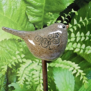 Op de afbeelding: Een keramiek vogelbeeldje met een bruine glazuur en een zwart bloemenmotief. De vogel zit op een metalen paal.