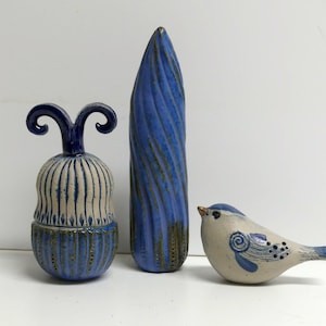 Op de afbeelding: Drie keramische sculpturen in blauw en wit. Een kleine vogel met blauwe accenten, een hoge, dunne, blauwe vaas en een kleine, ronde, blauw-witte pot met deksel.