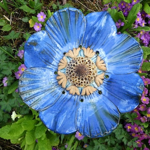 Op de afbeelding: Een grote blauwe keramische bloem met een getextureerd oppervlak en een bruin centrum. De bloem wordt omringd door paarse bloemen.