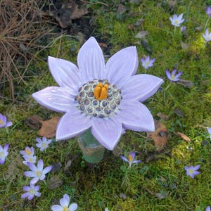 Op de afbeelding: Een grote, lichtpaarse keramische bloemensculptuur met een gedetailleerd midden, geplaatst in een tuin. De bloem heeft meerdere bloemblaadjes en is omgeven door kleine paarse krokussen en groen mos.