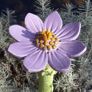 Op de afbeelding: Een keramische bloemensculptuur in lichtpaars, met zes bloemblaadjes en een geel centrum. De bloem staat tegen een achtergrond van groen en grijs blad. Een decoratief tuinelement.