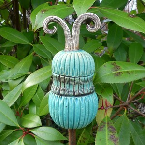Könnte beinhalten: Ein türkisblauer Keramik-Gartenpfahl mit einer weißen Spitze und zwei gekräuselten weißen Akzenten. Der Pfahl ist rostbraun und ragt aus dem Boden.