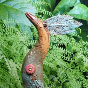 Op de afbeelding: Een handgemaakte keramische vogelsculptuur met een lange, gebogen nek en een gestileerde vleugel. De vogel heeft een bruin lichaam, een rode snavel en een blauwe vleugel. Een roze bloem siert het lichaam. De sculptuur staat tegen een achtergrond van groen gebladerte.