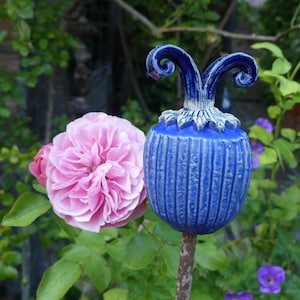 Op de afbeelding: Blauw keramisch tuinornament met een decoratieve top en verticale lijnen, naast een roze roos. Het ornament staat op een bruine stengel, tegen een achtergrond van groen gebladerte en paarse bloemen.