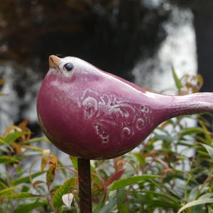 Puede incluir: Una escultura de pájaro de cerámica en tonos morados y blancos, posada sobre una varilla de metal. El pájaro tiene una superficie detallada y texturizada con un diseño floral en su costado. La escultura está situada sobre un fondo de follaje verde y un fondo borroso.