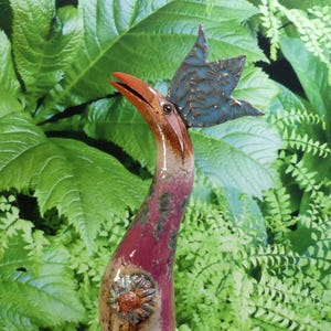 Op de afbeelding: Een keramische vogelsculptuur met een lange, gebogen nek. De vogel heeft een roodpaars lichaam, een bruin-oranje snavel en een blauwe, stervormige hoofdtooi. Een bloemversiering siert de borst. De achtergrond bestaat uit groene bladeren.