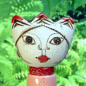 Può includere: Statuetta di gatto in ceramica con viso bianco, occhi marroni e dettagli rossi. Il gatto ha una corona e labbra rosse. La statuetta ha un corpo rosa e un colletto rosso. Lo sfondo è verde sfocato.