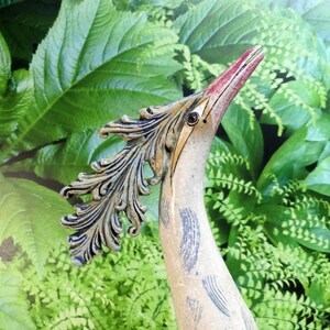 Op de afbeelding: Een keramische vogelsculptuur met een lange, gebogen nek en een gedetailleerde kop. De vogel heeft een rode snavel, een gedetailleerd oog en een decoratief veervormig element. De sculptuur is geschilderd in neutrale tinten met blauwe en bruine accenten, tegen een achtergrond van groen gebladerte.