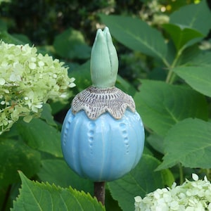 Op de afbeelding: Een lichtblauwe keramische tuinpaal met een bloemknopontwerp. De paal heeft een gedetailleerde, getextureerde kraag en een lichtgroene knop. Het blauwe gedeelte heeft een reeks kleine, verhoogde stippen. De paal staat tegen een achtergrond van groen gebladerte.