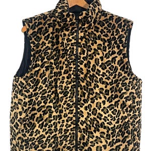Puede incluir: Un chaleco sin mangas con estampado de leopardo. El chaleco tiene un cuello alto y una cremallera negra de largo completo. El exterior presenta un estampado animal en marrón y beige, mientras que el interior es negro. El chaleco está colgado de una percha de madera.