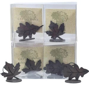 May include: Four sets of leaf-shaped place card holders in clear plastic boxes. Each set contains a dark brown metal leaf on a small base. The boxes have a beige background with a tree design and the words "CASUAL HOME place card".