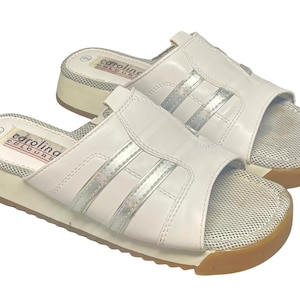 May include: A pair of white leather sandals with silver stripes and a mesh footbed. The sandals have a platform sole with a brown rubber bottom. The brand name "carolina colours" is printed on the insole.