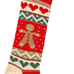May include: Red Christmas stocking with a knitted design. The stocking features a gingerbread man, hearts, and snowflakes. The name "Ella" is embroidered on the cuff. The toe and heel are cream-colored, and the cuff is red.