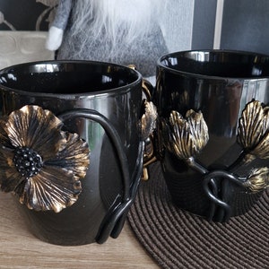 Könnte beinhalten: Zwei schwarze Keramikbecher mit goldenen Akzenten. Ein Becher hat einen goldenen Dollarzeichen-Griff und ein goldenes Blumendesign. Der andere Becher hat zwei goldene Blumendesigns.