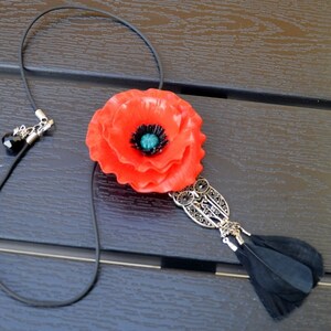 Op de afbeelding: Een ketting met een rode klaproos, een zwarte kwast en een zilveren hanger. De bloem is gemaakt van stof en heeft een groene kern.