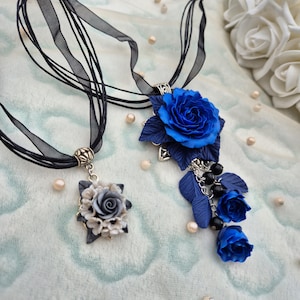 Op de afbeelding: Twee kettingen met zwarte lintkoorden. De ene hanger toont een tros grijze en witte rozen. De andere heeft een grote blauwe roos met donkerblauwe bladeren, een zilverkleurige metalen zetting en een bungelende ketting met blauwe rozenknoppen.
