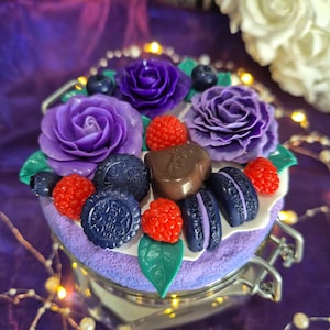 Peut inclure: Un pot décoratif avec un arrangement floral violet. L'arrangement comprend des roses violettes, des myrtilles, des framboises rouges et des biscuits bleu foncé. Le pot a un fermoir argenté et repose sur une surface violette.