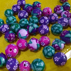 Könnte beinhalten: Eine Sammlung von lila, rosa und grünen Polymer-Tonperlen mit Blumen- und Blattmotiven. Die Perlen sind auf einer gelben Oberfläche angeordnet.