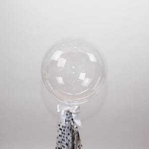 May include: A large, clear, round balloon with a white ribbon and a tassel of silver, black, and white confetti. The balloon is inflated and appears to be made of a transparent material. The balloon is a party decoration.
